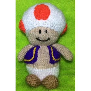 May include: A knitted mushroom character with a white body, orange cap, and purple vest. The character has a smiling face and is wearing a white diaper.