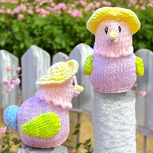 May include: Two knitted bird ornaments with yellow, pink, and green accents. The birds are sitting on white fence posts.