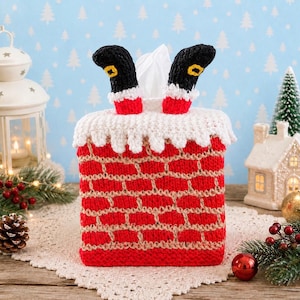 May include: A square tissue box cover crocheted to resemble a red brick chimney with a white snowy top. Two black boots with yellow buckles and white cuffs extend upward from the top, creating the appearance of legs disappearing into the chimney.