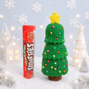 May include: A green crocheted Christmas tree with a yellow crocheted star on top and small colourful beads attached to the surface, standing next to a tall red rectangular box of Nestle Smarties Reindeer Drops sweets.
