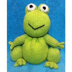 May include: A green knitted frog with white eyes and a black smile. The frog is sitting on a blue towel.