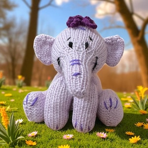 Könnte beinhalten: Ein gestricktes Lavendel-Elefanten-Spielzeug mit einer dunkelvioletten Schleife auf dem Kopf. Der Elefant hat große Ohren, einen Rüssel und violette Akzente an Füßen und Rüssel. Es sitzt auf grünem Gras mit kleinen Blumen.