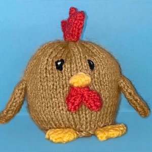 May include: A brown knitted chicken toy with red comb, yellow beak, and black eyes. The chicken has yellow feet and is sitting on a blue surface.