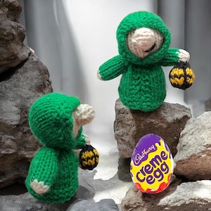 Puede incluir: Dos figuras verdes tejidas con capuchas y pequeñas linternas, de pie sobre rocas. También hay un huevo Cadbury Creme. Las figuras tienen caras blancas y sostienen linternas negras y amarillas.