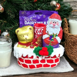 May include: A crocheted Christmas stocking filled with chocolate candy bars, including Cadbury Dairy Milk, Kit Kat, and a Santa Claus figure. The stocking is decorated with a red and white brick pattern and a green and red holly sprig.