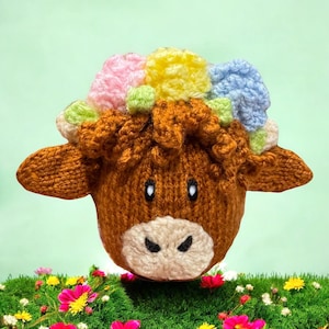 Op de afbeelding: Een handgemaakte bruin gebreide koeienkop met een kleurrijke, getextureerde manen. De koe heeft een witte snuit, zwarte ogen en kleine oren. De manen hebben roze, gele, groene en blauwe accenten. De koe staat tegen een lichtgroene achtergrond.