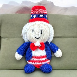 Puede incluir: Una muñeca de punto vestida como el Tío Sam. La muñeca lleva un sombrero rojo, blanco y azul, una chaqueta azul, una corbata de lazo roja y pantalones a rayas blancas y rojas. La muñeca tiene una cara blanca con ojos negros y una boca sonriente.