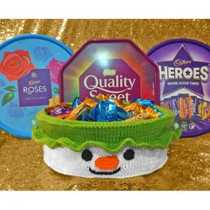 Puede incluir: Una cesta tejida en forma de muñeco de nieve llena de barras de chocolate surtidas. La cesta está sobre una superficie dorada brillante. Las barras de chocolate están en tres cajas diferentes: Cadbury Roses, Cadbury Heroes y Quality Street.