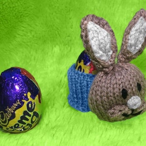 Puede incluir: Un porta huevos de conejo de punto marrón con ribete de punto azul. El conejo sostiene un huevo de chocolate envuelto en papel de aluminio morado y dorado.