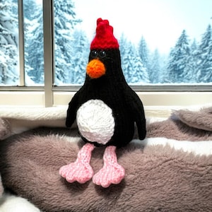 May include: A knitted penguin toy wearing a red knitted hat with black eyes and an orange beak. The penguin is sitting on a fluffy brown blanket.