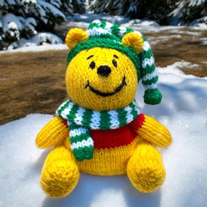 Könnte beinhalten: Ein gelber gestrickter Teddybär, der eine grün-weiß gestreifte Mütze und einen Schal trägt. Der Bär sitzt im Schnee.