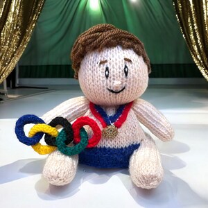 May include: A knitted doll wearing a blue and white swimsuit and a red, white, and blue ribbon with a gold medal. The doll is holding a set of five interlocking rings in red, yellow, blue, green, and black.