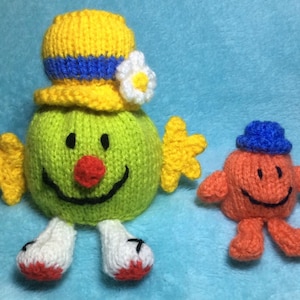 May include: Two knitted plush toys, one green with a yellow hat and a white flower, and one orange with a blue hat. Both toys have smiling faces.