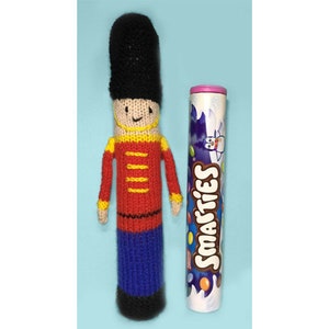 Puede incluir: Un soldado de juguete de punto con un uniforme rojo y amarillo y un sombrero negro. El soldado está de pie junto a un tubo de caramelos Smarties.