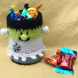 Può includere: Un porta caramelle di lana verde, grigio e nero a forma di mostro di Frankenstein con un viso sorridente. Il porta caramelle è pieno di caramelle al cioccolato assortite.