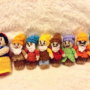 May include: Knitted dolls of Snow White and the Seven Dwarfs. Snow White is wearing a yellow dress and a red bow. The dwarfs are wearing different coloured hats and outfits.