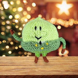 May include: A green knitted plush toy with a smiling face and brown legs. The toy is wearing a green knitted hat with a white trim.