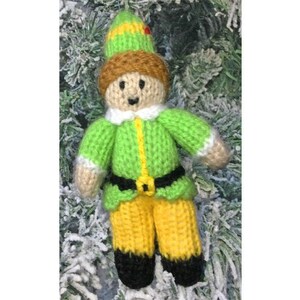 May include: A knitted elf ornament with a green and yellow outfit, brown hair, and a red hat. The elf is standing on a snowy background.