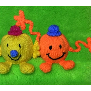 May include: Two knitted plush toys in the shape of smiling faces. The first toy is yellow with a pink hat and blue eyes. The second toy is orange with a blue hat and black eyes. Both toys have a knitted smile and a knitted nose.