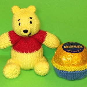 May include: A knitted Winnie the Pooh bear wearing a red shirt sits next to a knitted orange chocolate egg. The egg wrapper says "Made with real orange oil".