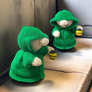 Könnte beinhalten: Zwei gestrickte Figuren mit grünen Kapuzenmänteln, die gelb-schwarze Laternen halten. Die Figuren haben weiße Gesichter und Hände. Sie stehen auf einer Betonoberfläche.