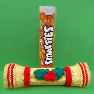Puede incluir: Un cracker de Navidad tejido con rayas rojas y blancas y una rama de acebo verde con bayas rojas. El cracker está al lado de una caja de caramelos Smarties naranja.