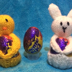 May include: Two knitted Easter egg holders, one shaped like a chick and the other like a bunny, each holding a purple foil-wrapped chocolate egg. The eggs are labeled "Cadbury Creme Egg".