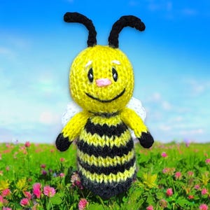 Puede incluir: Un juguete de abeja de punto amarillo y negro con una cara sonriente y alas blancas. La abeja está de pie en un campo de hierba verde y flores rosas.