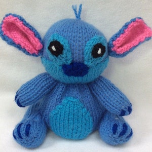 May include: A blue knitted Stitch plush toy with pink ears and black eyes. The toy is sitting on a white surface.