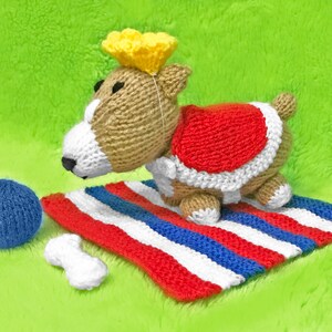 May include: A knitted dog toy wearing a red and white crown. The dog is laying on a red, white, and blue striped blanket with a blue ball and a white bone.