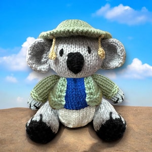 May include: A grey knitted koala wearing a green hat, a green jacket, and a blue shirt. The koala is sitting on a brown surface.