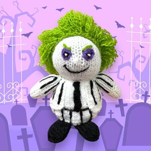 May include: A knitted plush toy of a character with green hair, a black and white striped suit, and a mischievous grin. The toy is standing in front of a purple background with a graveyard scene.
