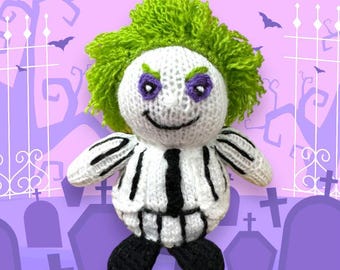 May include: A knitted plush toy of a character with green hair, a black and white striped suit, and a mischievous grin. The toy is standing in front of a purple background with a graveyard scene.
