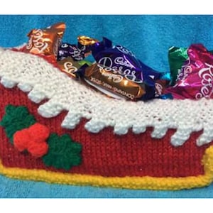Könnte beinhalten: Ein gestrickter roter und weißer Weihnachts-Schlitten gefüllt mit verschiedenen Schokoladenbonbons in Folie verpackt. Der Schlitten hat einen grünen Stechpalmenzweig mit einer roten Beere an der Seite.
