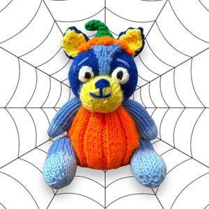 May include: A blue and yellow knitted plush toy dressed as a pumpkin for Halloween. The toy has a green stem on its head and a large orange pumpkin body.