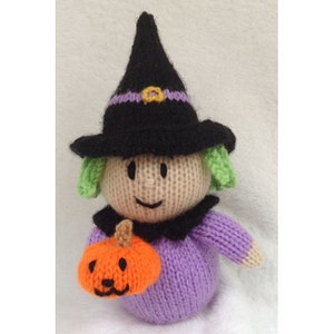 May include: A knitted witch doll wearing a black witch hat with a purple band and holding an orange pumpkin. The doll has a purple dress, green hair, and a smiling face.