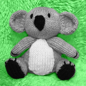 May include: A grey knitted koala plush toy with black eyes and a white tummy. The toy is sitting on a green fluffy surface.