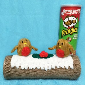 May include: A knitted yule log with two knitted robins perched on top. The yule log is decorated with red holly berries. A can of Pringles Sour Cream & Onion crisps is in the background. The can has a cartoon character with a moustache and bow tie on it. The text on the can reads "Free Present with Every Can" and "Pringles".