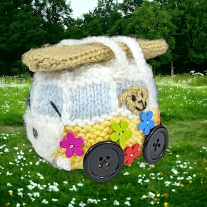 May include: A handmade, knitted camper van in white, yellow, and blue, with a tan roof and handle. The van features black button wheels and colorful flower accents. A small, tan dog is in the window. Set against a green field.