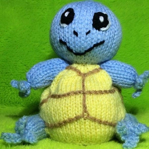 May include: A handmade knitted blue turtle plush toy with a yellow shell and brown accents. The turtle has black eyes and a smiling mouth. The toy is sitting on a green background.