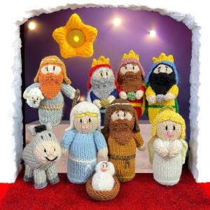 Könnte beinhalten: Eine gestrickte Krippenszene mit Figuren der Heiligen Drei Könige, Maria, Josef, Jesuskind, einem Engel und einem Esel. Die Figuren haben farbenfrohe Kleidung und sind vor einem lila Hintergrund mit einem Stern und Lichtern platziert.