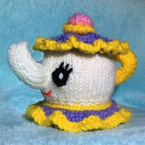 May include: A knitted teapot with a yellow, purple, and white ruffle trim. The teapot has a pink lid and a black eye with a white pupil.