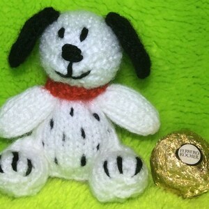 May include: A white knitted dalmatian dog toy with black spots and a red collar. The toy is sitting on a green fluffy surface next to a gold wrapped chocolate.