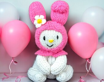 Könnte beinhalten: Ein rosa und weiß gestricktes Hasenspielzeug mit einer Blume auf dem Kopf. Der Hase sitzt auf einer weißen Oberfläche, umgeben von rosa und weißen Luftballons.