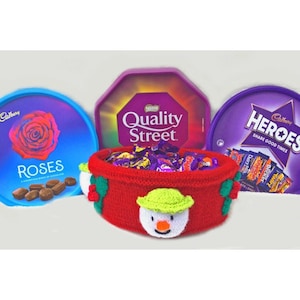 Könnte beinhalten: Ein roter gestrickter Schneemann-Korb gefüllt mit verschiedenen Cadbury-Schokoladentafeln. Der Korb ist umgeben von drei runden Cadbury-Schokoladenboxen: Roses, Quality Street und Heroes. Die Boxen sind lila, blau und grün.