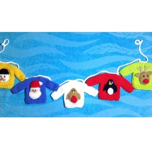 May include: Five miniature knitted Christmas jumper decorations. Each jumper is a different colour, including yellow, blue, white, red, and green, and features a different festive design, such as a snowman, Santa, penguin, and reindeer.