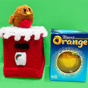Könnte beinhalten: Ein gestrickter roter Briefkasten mit einer weißen, schneebedeckten Oberseite und einem kleinen gestrickten Rotkehlchen, das darauf sitzt. Daneben eine Schachtel Terry's Chocolate Orange, mit blauem Hintergrund und orangefarbener Grafik.