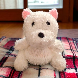 May include: A white plush dog toy with pink inner ears, black eyes, and a black nose. The toy is sitting on a plaid blanket. The dog has a knitted chest and fluffy fur.