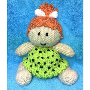 May include: A knitted doll with orange hair, a white headband, and a green polka dot dress. The doll is sitting on a blue towel.