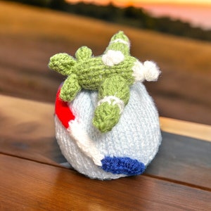 May include: A handmade knitted airplane toy in green and white, resting on a light blue knitted ball. The ball has red, white, and blue accents. The toy is likely a children's item or a decorative piece.